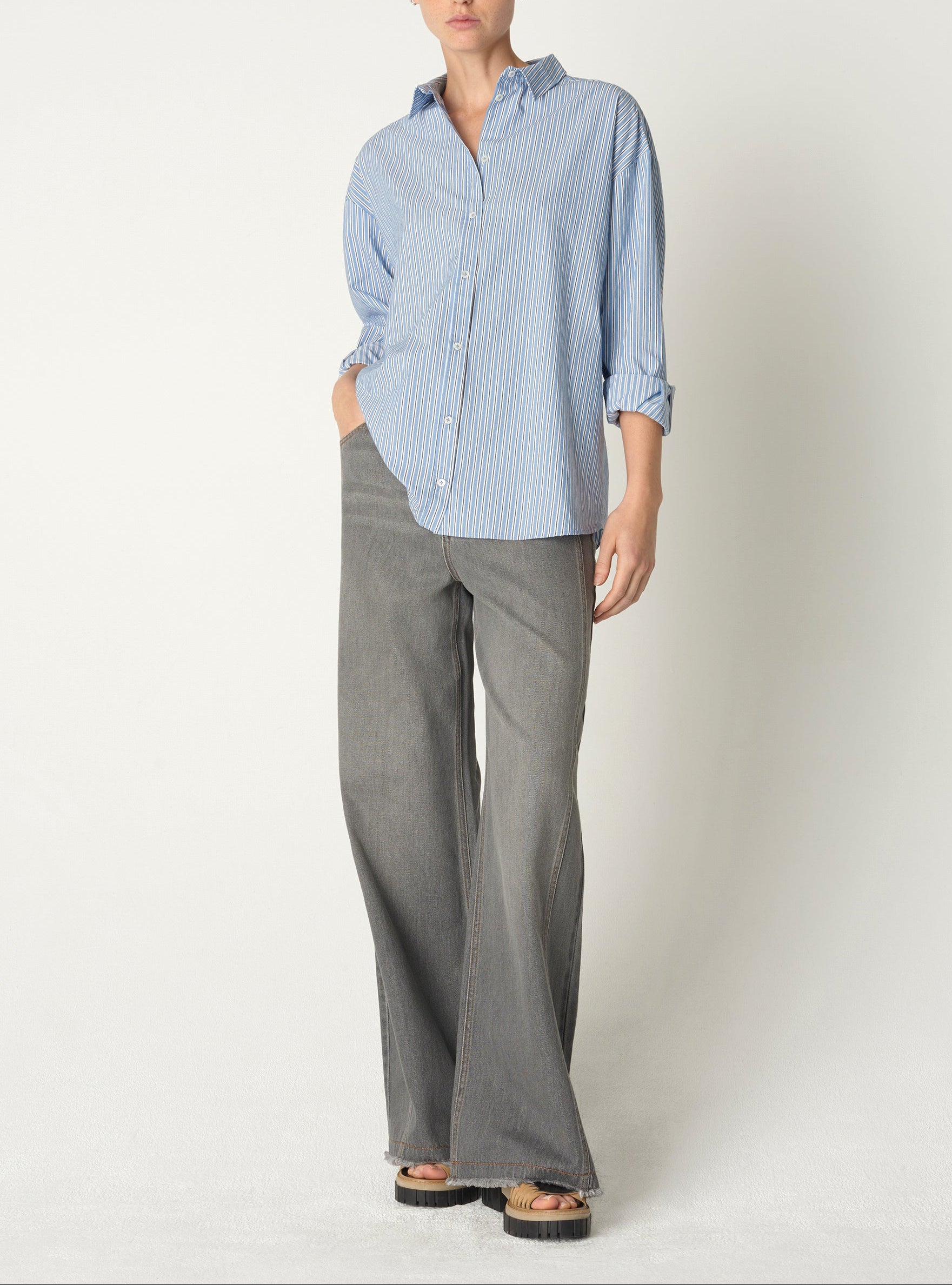 Une personne portant une chemise bleue rayée et un pantalon gris se tient debout sur un fond blanc uni.